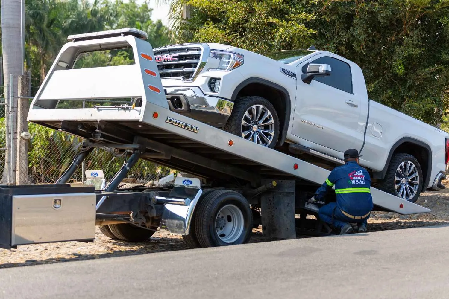 A white pickup truck is being loaded onto a flatbed tow truck by a worker outdoors, near a road lined with trees.