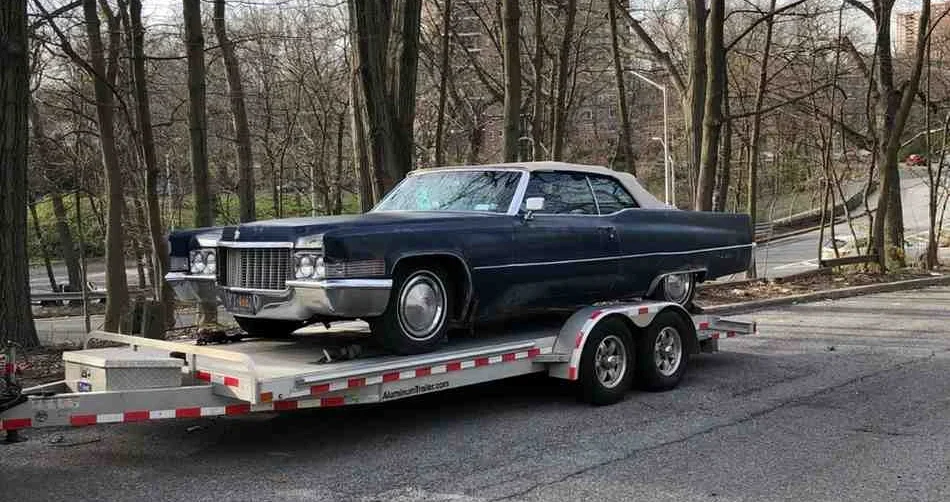 A classic car being towed on a flatbed tow truck parked near a wooded area.