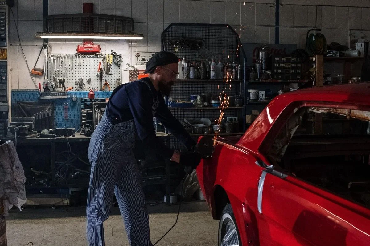 Mechanic restoring a vintage red car in a well-equipped garage with sparking grinder.