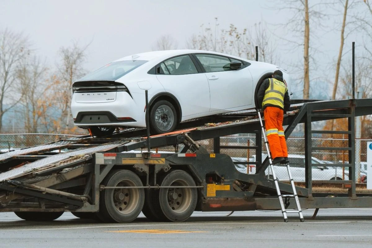 A worker in reflective gear loading a white car onto a flatbed truck ramp.