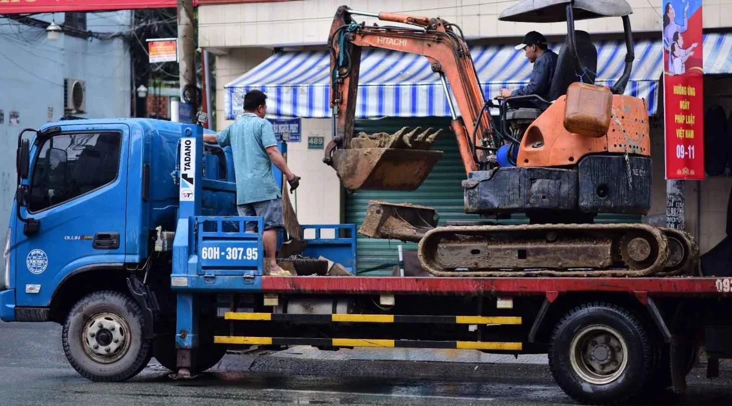 An orange backhoe loader sitting on a flatbed trailer in a market yard.