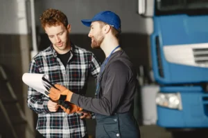 Mechanic in uniform consults with customer in garage setting beside a blue truck.