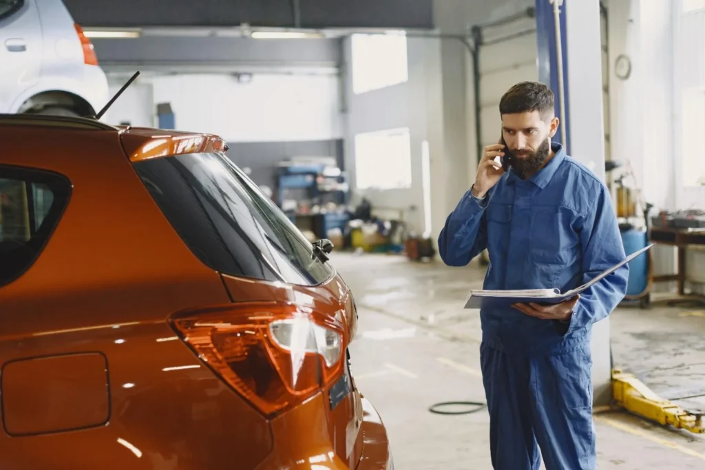 A mechanic in a blue overall with papers in his hand is making a phone call