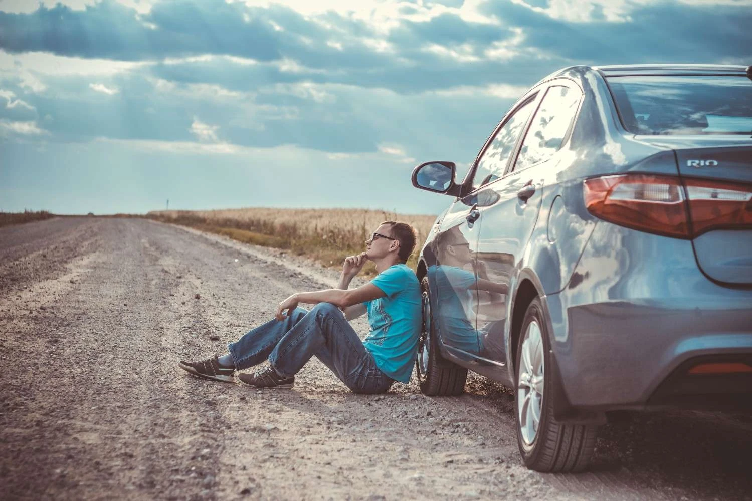 A person sitting on the ground next to a silver car on a gravel road.