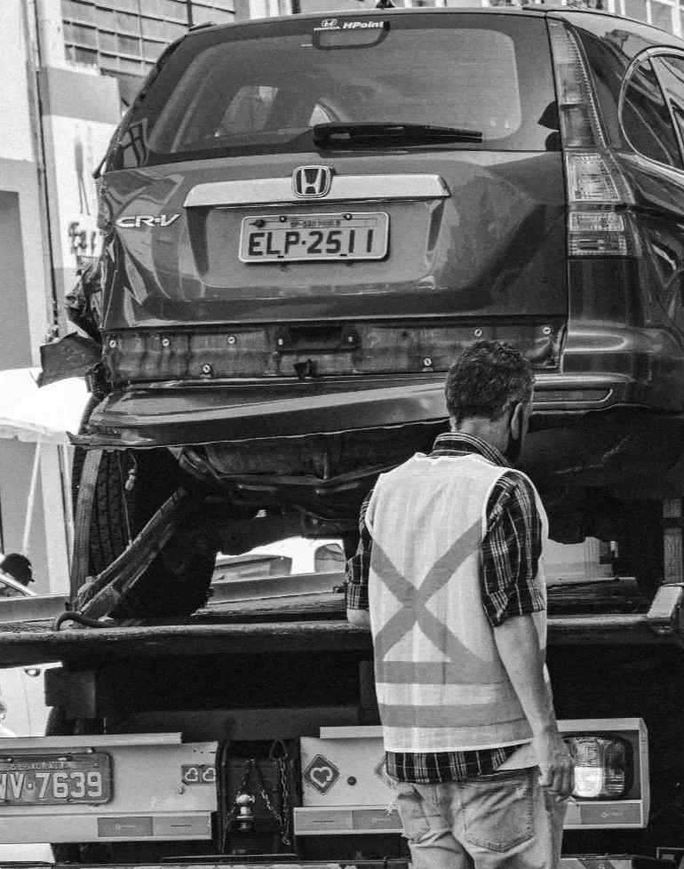 A man wearing a safety vest watches as a tow truck lifts a Honda Car onto its platform in a city street.