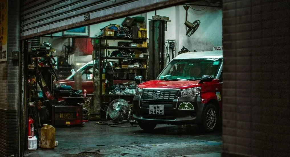 Red van inside an auto repair shop with open roller shutter, showcasing tools and equipment.